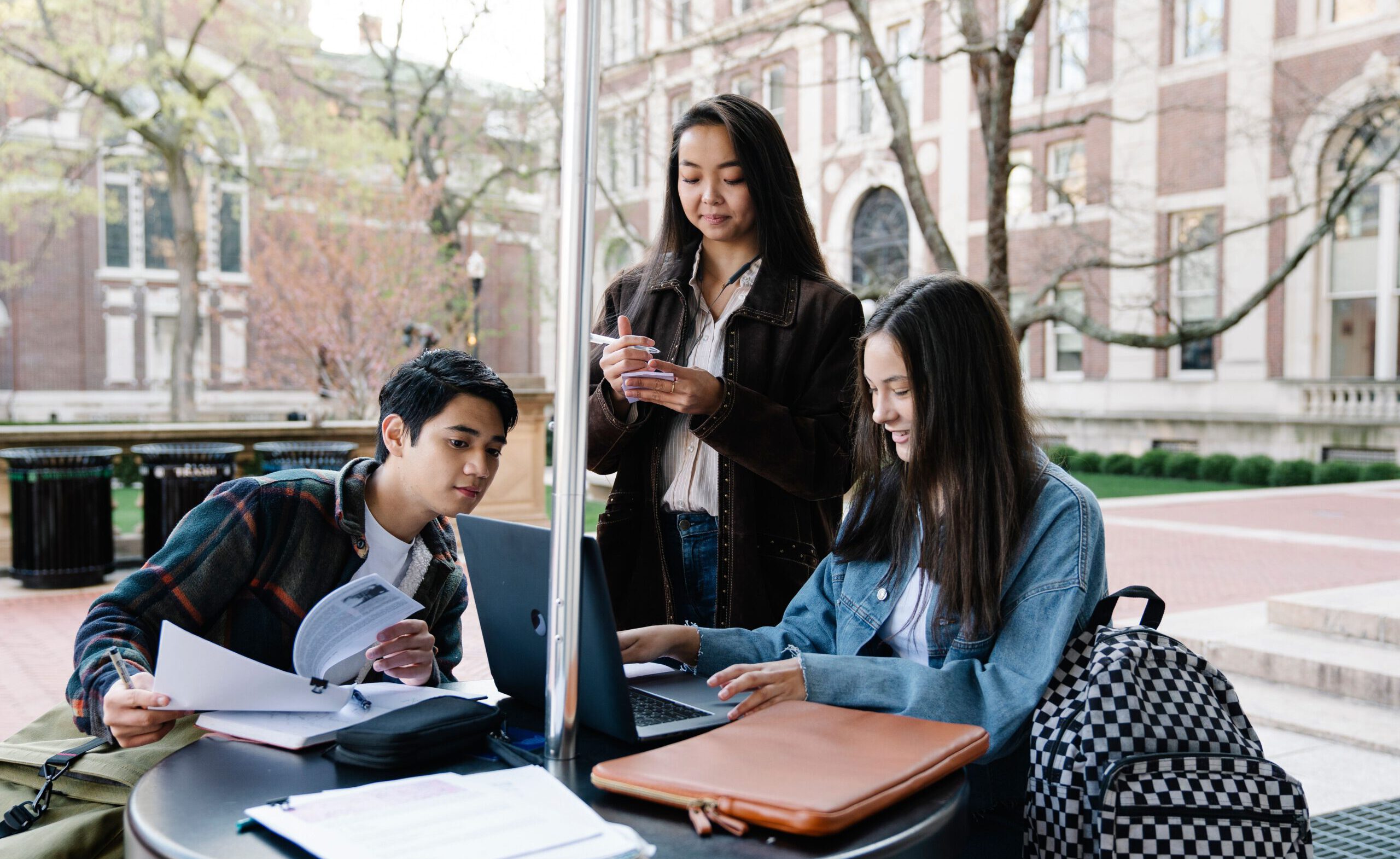 Students collaborate on a project outside a university building, engaged with their devices and materials.